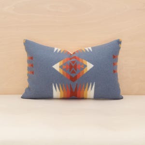 May include: A rectangular, blue cushion with a geometric pattern in orange, white, and yellow. The cushion has a Southwestern design, with a central diamond shape and repeating patterns along the sides. The cushion is on a white surface.