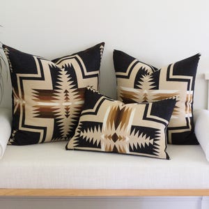May include: Three scatter cushions featuring a geometric pattern in black, beige, and brown. Two cushions are square-shaped, and one is rectangular. The design consists of sharp, angular lines and serrated edges arranged in a symmetrical, repeating motif.