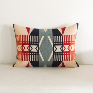 May include: Rectangular cushion with a geometric pattern in red, navy, cream, and grey. The cushion features a symmetrical design with vertical stripes and diamond shapes. The edges are trimmed with navy blue fabric.