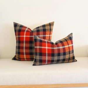 May include: Two plaid throw pillows in shades of red, orange, navy, and tan. The pillows are square and have a classic plaid pattern. They are placed on a white bench.