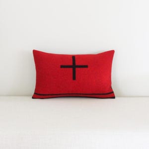 May include: A rectangular red pillow with a black cross design. The pillow is placed on a white surface. The pillow has a black border.