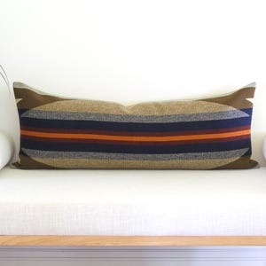 May include: A long rectangular lumbar cushion featuring a woven pattern of horizontal stripes in shades of olive green, dark blue, grey, and orange. The design includes a large, centred oval shape that spans the length of the cushion cover.