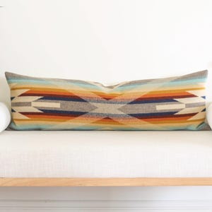 May include: Rectangular decorative pillow with a Southwestern geometric pattern in shades of blue, orange, and beige. The pillow is placed on a white bench with two white bolster pillows on each side. The pillow's design features a symmetrical arrangement of shapes and colors.