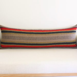 K&ouml;nnte beinhalten: Ein langes, rechteckiges Kissen mit einem Streifenmuster in Braun-, Rot-, Schwarz- und Gelbt&ouml;nen. Das Kissen liegt auf einem wei&szlig;en Sofa. Das Kissen ist ca. 30 cm breit und 91 cm lang.