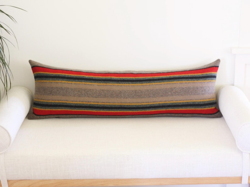 K&ouml;nnte beinhalten: Ein langes, rechteckiges Kissen mit einem Streifenmuster in Braun-, Rot-, Schwarz- und Gelbt&ouml;nen. Das Kissen liegt auf einem wei&szlig;en Sofa. Das Kissen ist ca. 30 cm breit und 91 cm lang.