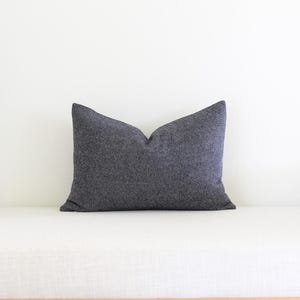 May include: A dark gray decorative pillow with a textured herringbone pattern. The pillow is rectangular and rests on a white bench. The pillow is a neutral color and would complement many home decor styles.