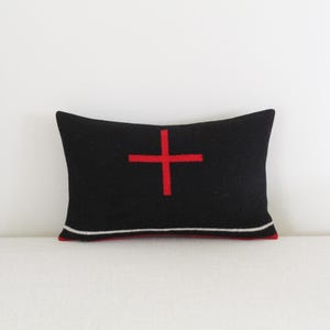 May include: A rectangular black pillow with a bold red cross design in the center. The pillow has a red and white trim along the bottom edge. The pillow is a decorative home accent.