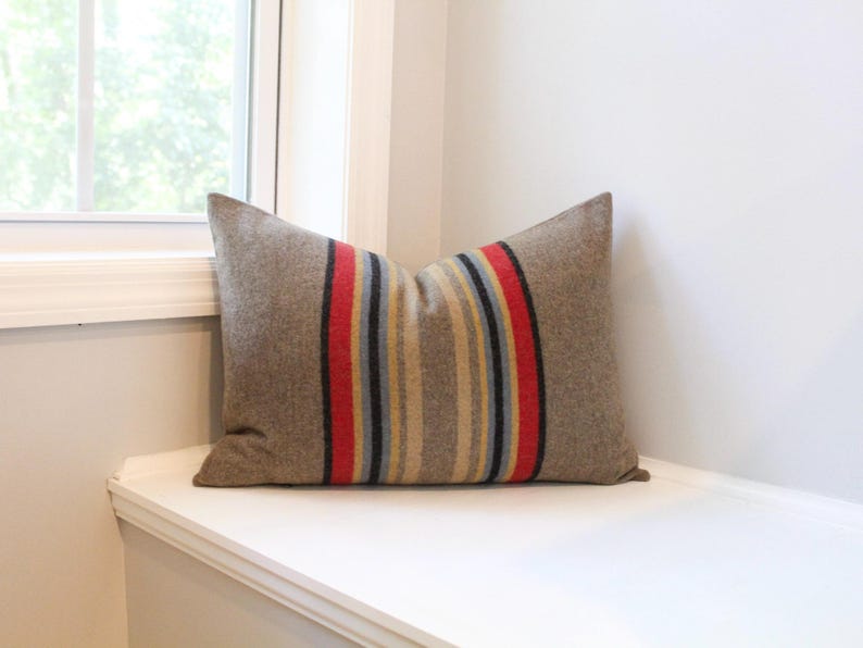 May include: A rectangular decorative pillow with a brown background and vertical stripes of red, yellow, black, and blue. The pillow is resting on a white windowsill, near a window with a view of trees.