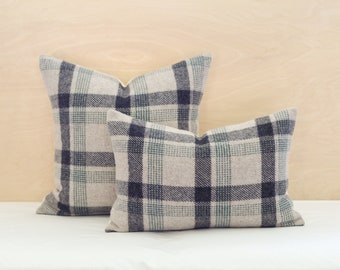 May include: Two cushions with a plaid pattern in shades of grey, navy, and teal. The cushions are rectangular and square, against a neutral background.