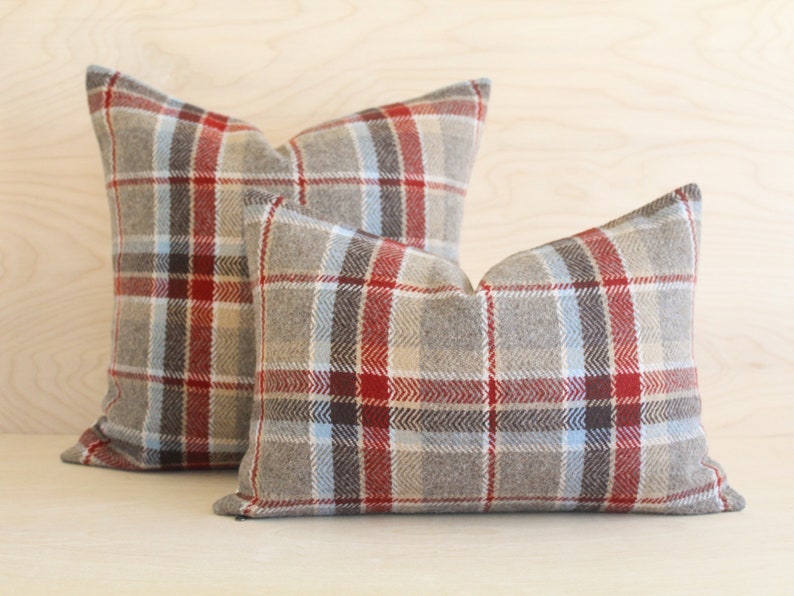 Op de afbeelding: Twee plaid kussens met een rood, blauw en bruin patroon op een lichte houten achtergrond.