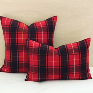 May include: Two red and black plaid cushions. The cushions feature a traditional tartan design with black, red, yellow, and white stripes. The cushions are rectangular and square, and are placed on a white surface.