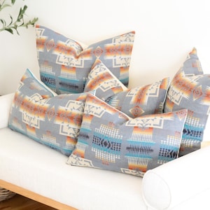 May include: A collection of patterned throw cushions in shades of grey, orange, blue, and cream, arranged on a white sofa. The cushions feature a geometric design, adding a touch of warmth and texture to the room. The sofa is white.