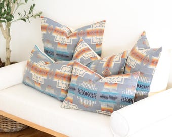 May include: Several decorative throw pillows with a Southwestern geometric pattern in shades of gray, orange, blue, and cream are arranged on a white sofa. The pillows have a soft, textured appearance, adding a cozy touch to the living space.