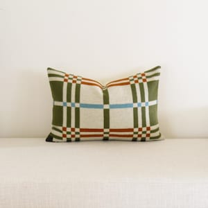 May include: Rectangular decorative pillow with a plaid pattern in olive green, cream, rust, and light blue. The pillow is placed on a white bench. The pillow's design features a grid-like arrangement of intersecting lines and blocks of color.