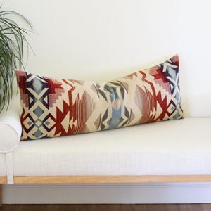 May include: A long rectangular lumbar pillow featuring a woven pattern of geometric shapes in shades of deep red, cream, light blue, and dark brown. The pillow has a textured fabric surface and is positioned horizontally on a white bench seat.