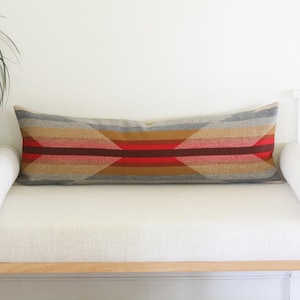 May include: A long rectangular lumbar pillow featuring a woven geometric pattern in horizontal stripes of gray, tan, brown, and vibrant red. The pillow has a textured fabric finish and is centered on a white bench with two white bolster pillows.