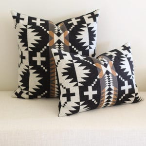 May include: Two square throw cushions with a geometric pattern in black, white, brown, and grey. The cushions are placed on a white surface. The design features crosses and zigzags.