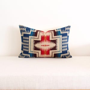 May include: Rectangular cushion with a geometric pattern in shades of blue, red, and beige. The cushion features a central design with a star-like shape and is placed on a white surface.