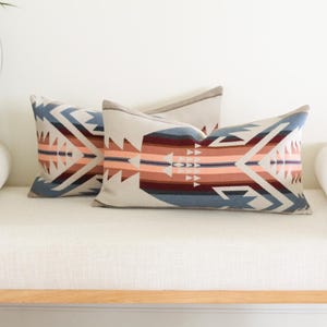 May include: Three rectangular lumbar cushions with a Southwestern geometric design in blue, peach, and burgundy. They rest on a white bench with a wooden frame. The cushions are approximately 60cm long.