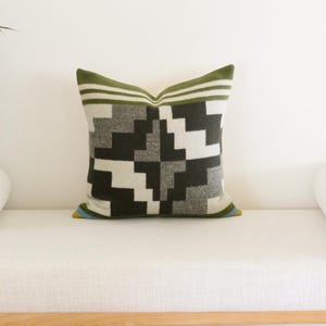 May include: Square decorative pillow with a geometric pattern in shades of black, gray, and white. The pillow has a green border with white stripes and is placed on a white bench. The pillow is approximately 18 inches square.