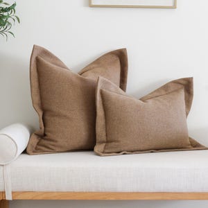 May include: Two brown throw pillows with a textured fabric on a white linen bench with a white bolster pillow.