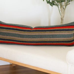 May include: A long, rectangular pillow with a striped pattern in shades of gray, red, yellow, and black. The pillow is made of a soft, woven fabric and is perfect for adding a touch of color and texture to any room.
