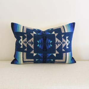 May include: Rectangular decorative pillow with a geometric pattern in shades of blue, gray, and white. The pillow has a dark blue background with a symmetrical design. The pillow is placed on a white surface.