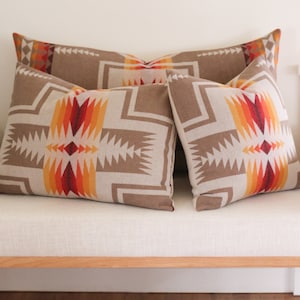 May include: Three decorative throw pillows with a geometric pattern in shades of brown, orange, and red. The pillows are arranged on a white linen sofa.