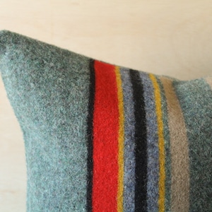 May include: A green, gray, black, red, and yellow striped throw pillow made of wool. The pillow is a decorative accent for a home.