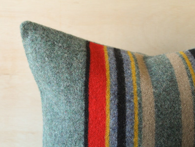 May include: A green, gray, black, red, and yellow striped throw pillow made of wool. The pillow is a decorative accent for a home.