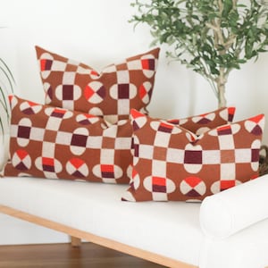 May include: Three brown and white patterned throw pillows with red and burgundy accents are arranged on a white bench with a white bolster pillow. The pillows have a geometric pattern of squares and circles.