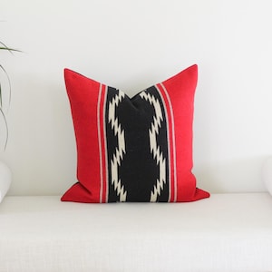 May include: A decorative pillow with a red border and a central black and white geometric pattern. The pillow is square-shaped and rests on a white surface, offering a pop of color to any room. The pillow is a decorative home accent.