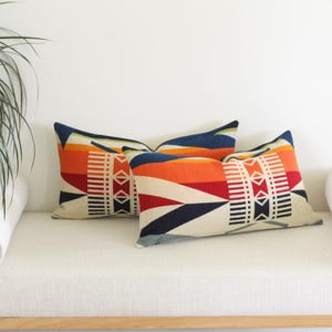 May include: Two rectangular cushions with a geometric pattern in shades of orange, red, navy blue, and cream. The cushions are placed on a white bench. The design features vertical and horizontal lines and diamond shapes.