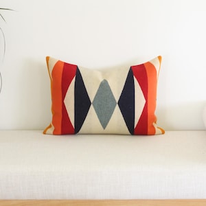 May include: A rectangular cushion with a geometric pattern. The design includes a cream-coloured base with a central diamond pattern in navy and grey, and red and orange stripes. The edges are trimmed in orange. Measures approximately 40cm x 60cm.