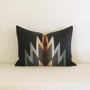 May include: A rectangular throw pillow featuring a dark charcoal gray base with a central geometric pattern. The design consists of symmetrical, jagged, mountain-like peaks in shades of cream, light blue, mustard yellow, and deep brown.