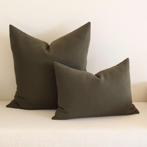 May include: Two olive green throw pillows of different sizes rest on a white surface. The larger pillow is upright, while the smaller one is angled. The pillows have a simple, solid design, suitable for home decor.