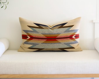 May include: A rectangular throw cushion featuring a beige base colour with a prominent geometric pattern in shades of blue, brown, red, and tan. The design consists of symmetrical, angular shapes radiating from a central horizontal red stripe.