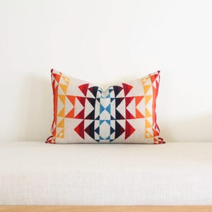 May include: A rectangular cushion featuring a geometric pattern in red, orange, yellow, blue, and white. The cushion is placed on a white sofa, highlighting its colourful design and texture.