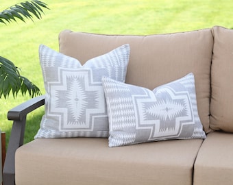 May include: Two decorative throw pillows with a geometric pattern in gray and white rest on a beige outdoor sofa. The pillows feature a central cross-like design with intricate details, adding a touch of Southwestern style to the outdoor setting.