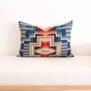 May include: Rectangular cushion with a geometric pattern in shades of blue, red, brown, and cream. The cushion features a Southwestern-inspired design, adding colour and pattern to any living space.