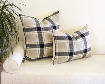 May include: Two square cushions with a beige, black, and white plaid design. They are placed on a white bench. A green plant is visible in the background. The cushions would be suitable for a living room.
