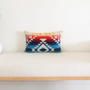 May include: Rectangular cushion featuring a geometric design in red, orange, blue, and white. The cushion is placed on a white bench with cylindrical bolsters. The cushion measures approximately 30 cm by 50 cm.