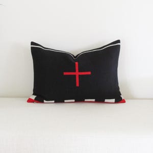 May include: A rectangular black pillow with a red cross design in the center. The pillow has a white border and red and white striped accents on the bottom. The pillow is approximately 20 inches long.