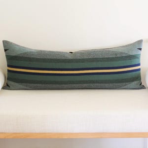 May include: A long, rectangular cushion with grey, green, and yellow horizontal stripes. It rests on a white bench, flanked by white bolster cushions. The cushion's ends feature a geometric design. The bench is made of wood.