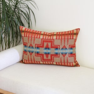 May include: Rectangular decorative pillow with a geometric pattern in red, tan, and blue. The pillow is resting on a white bench next to a white bolster pillow and a green plant.