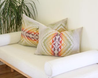 May include: Two decorative throw pillows with a geometric pattern in shades of green, orange, and purple. The pillows are on a white daybed with white bolster pillows. The design has a Southwestern or Native American aesthetic.