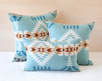 May include: Three decorative throw pillows featuring a geometric pattern in shades of blue, brown, and white. The pillows are arranged in a pyramid shape, with the largest pillow at the bottom and the smallest pillow at the top.