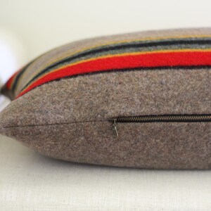 K&ouml;nnte beinhalten: Ein rechteckiges Kissen mit braunem Wollbezug. Das Kissen hat horizontale Streifen in Rot, Gelb, Schwarz und Gold. Ein Rei&szlig;verschluss verl&auml;uft an der Seite. Das Kissen liegt auf einer wei&szlig;en Oberfl&auml;che.