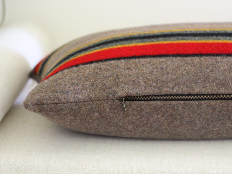 K&ouml;nnte beinhalten: Ein rechteckiges Kissen mit braunem Wollbezug. Das Kissen hat horizontale Streifen in Rot, Gelb, Schwarz und Gold. Ein Rei&szlig;verschluss verl&auml;uft an der Seite. Das Kissen liegt auf einer wei&szlig;en Oberfl&auml;che.