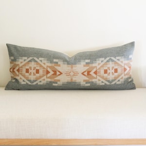 May include: A long rectangular lumbar pillow featuring a gray base color with a repeating geometric pattern in shades of terracotta, cream, and light gray. The design consists of symmetrical diamond shapes and linear motifs centered across the fabric.